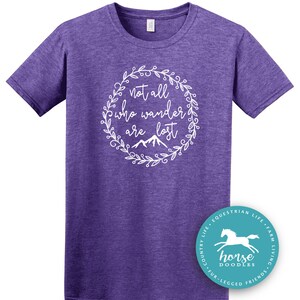 以下が含まれることがあります： 白い花のリースデザインで「not all who wander are lost」というテキストが入った紫色のTシャツ。右下には馬のグラフィックが入った丸いロゴがあります。このシャツは杢紫です。
