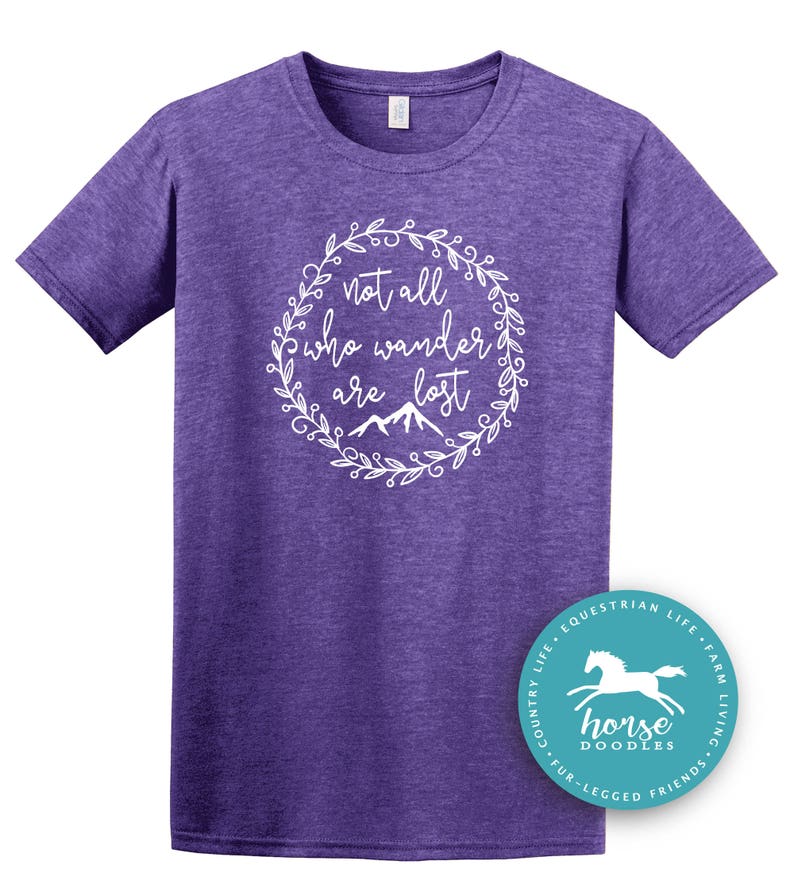 以下が含まれることがあります： 白い花のリースデザインで「not all who wander are lost」というテキストが入った紫色のTシャツ。右下には馬のグラフィックが入った丸いロゴがあります。このシャツは杢紫です。