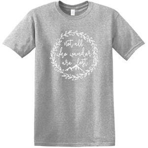 以下が含まれることがあります： 白の円形デザインのグレーのTシャツ。デザインは、筆記体のフォントで「Not all who wander are lost」というテキストを特徴とし、花のリースと山のグラフィックに囲まれています。シャツは半袖で、クルーネックです。