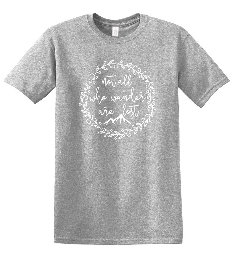 以下が含まれることがあります： 白の円形デザインのグレーのTシャツ。デザインは、筆記体のフォントで「Not all who wander are lost」というテキストを特徴とし、花のリースと山のグラフィックに囲まれています。シャツは半袖で、クルーネックです。