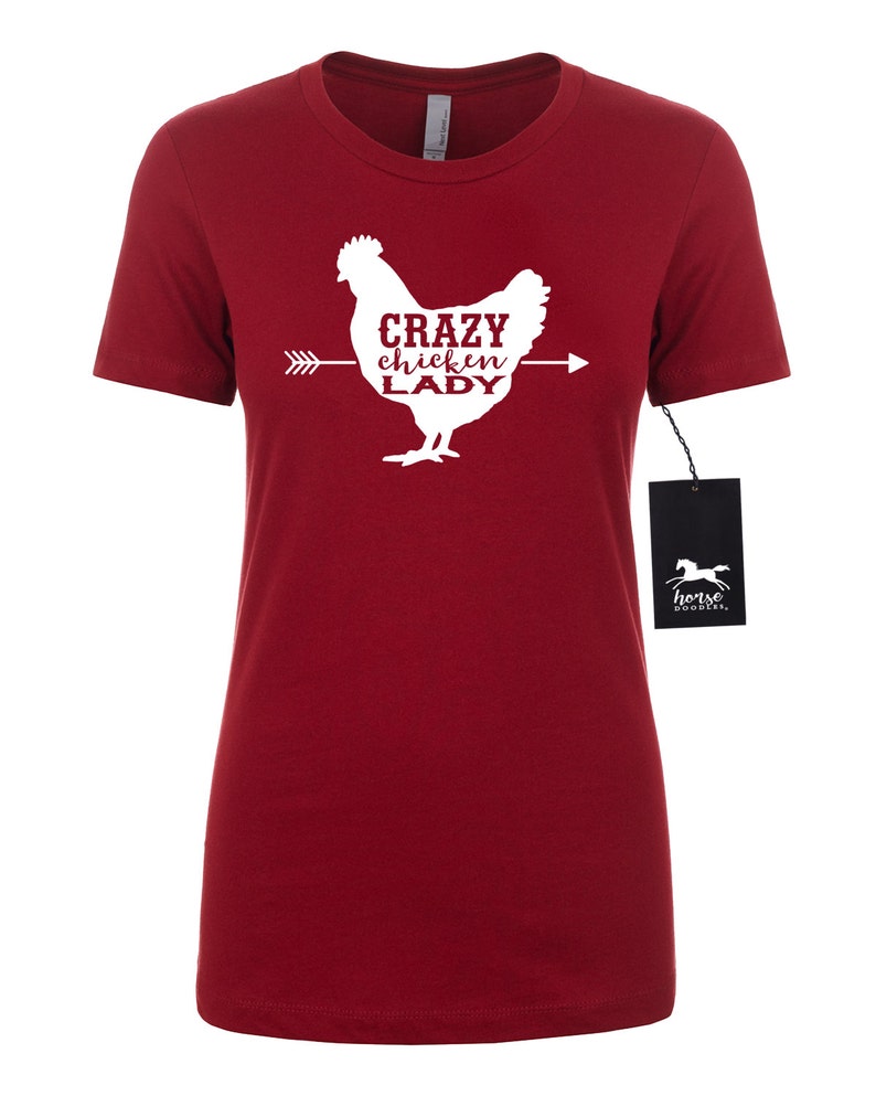 May include: A red t-shirt featuring a white graphic of a chicken with the text "CRAZY chicken LADY" in a serif font. An arrow points from left to right through the chicken. A black tag with a horse logo hangs from the shirt.