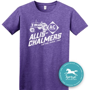 May include: Purple t-shirt with a white graphic of a tractor and the words "ALLIS-CHALMERS ESTABLISHED 1901". A round sticker with a horse graphic is on the bottom right. The shirt is a crew neck.