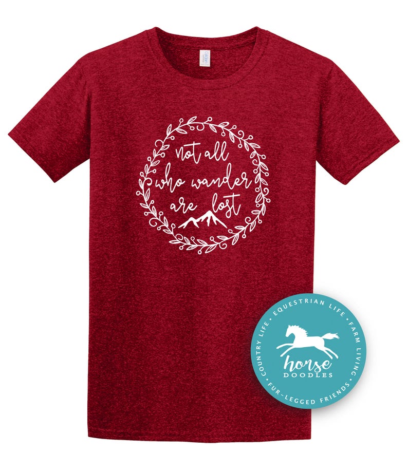以下が含まれることがあります： 白い花のリースの中に「not all who wander are lost」というテキストが入った、マルーン色のTシャツ。馬のグラフィックと「horse doodles」という言葉が入った円形のロゴが右下にあります。
