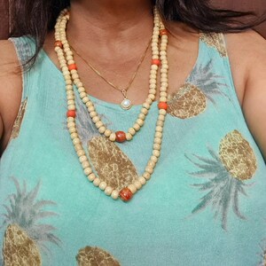 May include: A layered necklace set featuring a gold chain with a small pendant and two beaded necklaces. The longer necklaces have light-colored wooden beads interspersed with orange and reddish-brown beads. The background is a light blue top with a pineapple pattern.