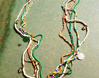 May include: A multi-strand necklace with colorful beads in white, green, red, orange, yellow, and blue. The necklace features small charms and a white tassel pendant. The jewelry is displayed on a green surface.