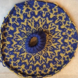 Puede incluir: Un artículo circular tejido en azul y amarillo. El diseño presenta un patrón geométrico repetitivo. El centro tiene un círculo azul más pequeño y elevado. Los bordes están acabados con un ribete amarillo. El artículo parece hecho a mano.
