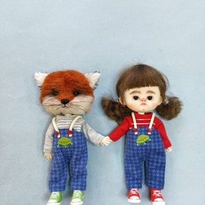 May include: Two plush toys, a fox and a girl, are wearing blue overalls with a green turtle design. The fox has a red and white striped shirt and the girl has a red shirt with white stripes. Both toys are holding hands.