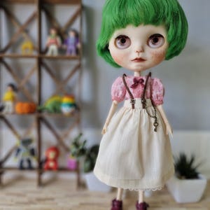 Puede incluir: Una muñeca con el pelo verde que lleva un vestido rosa y blanco con tirantes marrones y un charm de llave. La muñeca tiene ojos marrones y un rubor rosa.