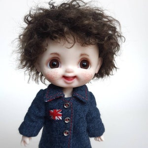 May include: A small doll with brown curly hair wearing a blue denim jacket with a red embroidered Union Jack patch. The doll has a smiling face with rosy cheeks and brown eyes.