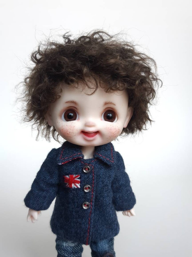 May include: A small doll with brown curly hair wearing a blue denim jacket with a red embroidered Union Jack patch. The doll has a smiling face with rosy cheeks and brown eyes.