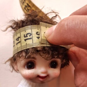 May include: A close-up of a doll's head with brown curly hair being measured with a yellow tape measure. The tape measure reads 15.5 cm = 6 ins.
