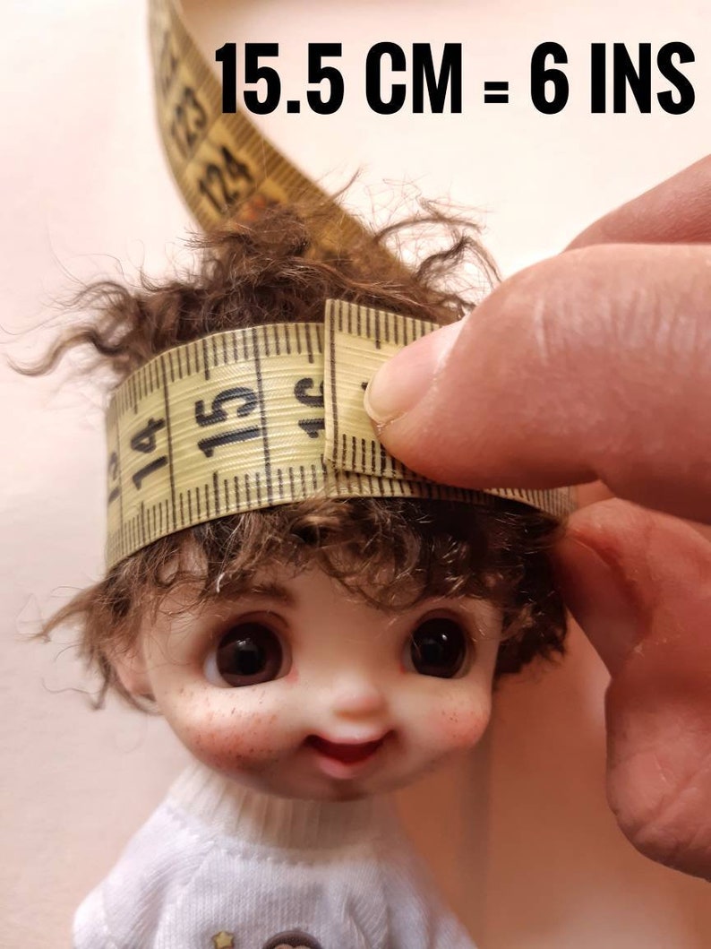 May include: A close-up of a doll's head with brown curly hair being measured with a yellow tape measure. The tape measure reads 15.5 cm = 6 ins.
