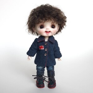 May include: A small doll with brown curly hair wearing a blue denim jacket with a red, white and blue patch, blue jeans, and black shoes with red laces.