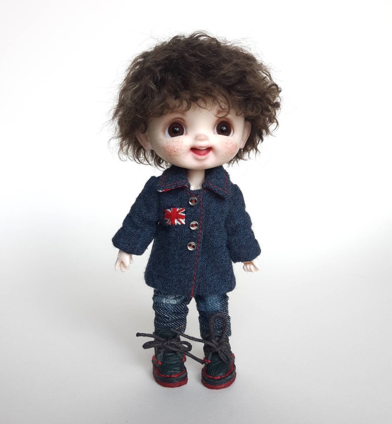 May include: A small doll with brown curly hair wearing a blue denim jacket with a red, white and blue patch, blue jeans, and black shoes with red laces.