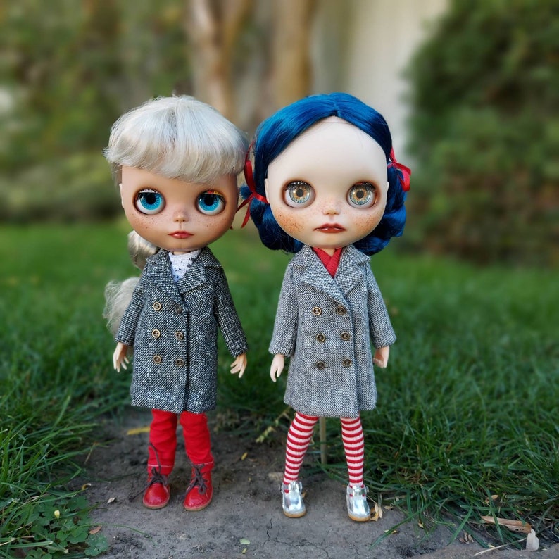 May include: Two dolls wearing gray coats with red and white striped pants and red shoes. The doll on the left has blonde hair and blue eyes. The doll on the right has blue hair and blue eyes.