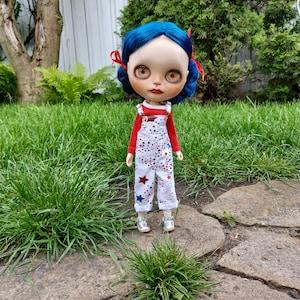 Puede incluir: Una muñeca con el pelo azul que lleva un mono estampado con estrellas rojas y blancas. La muñeca está de pie en un camino con hierba verde.