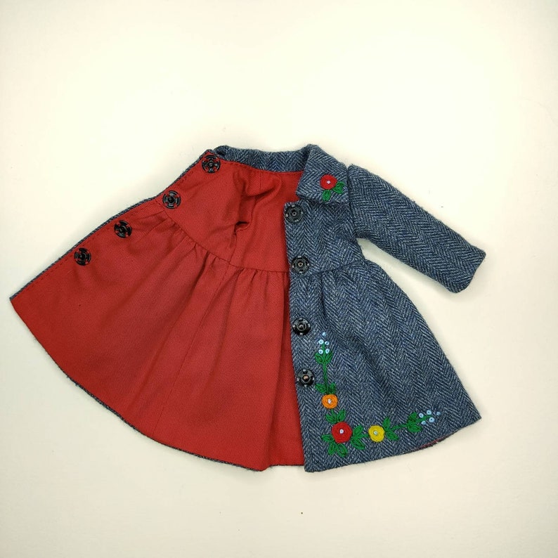 Puede incluir: Un abrigo reversible azul y rojo con un dise&ntilde;o de bordado floral. El abrigo tiene cierre de botones y un dobladillo con volantes.