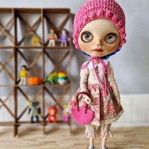 Puede incluir: Una muñeca con el pelo azul que lleva un gorro y un vestido de crochet rosa, sosteniendo un bolso de crochet rosa. La muñeca está de pie sobre un suelo de madera.