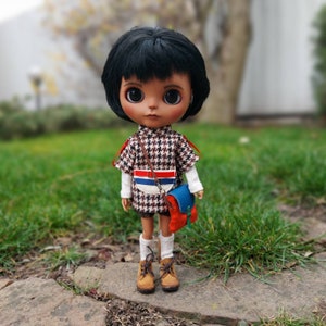 Puede incluir: Una muñeca con cabello negro y ojos marrones que lleva un vestido a cuadros marrones y blancos con una franja azul y roja, una correa marrón y una pequeña bolsa roja y azul. La muñeca lleva botas marrones.