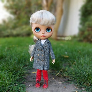 May include: A doll with blonde hair wearing a gray tweed coat, red pants, and red shoes. The doll has blue eyes and is standing on a grassy surface.