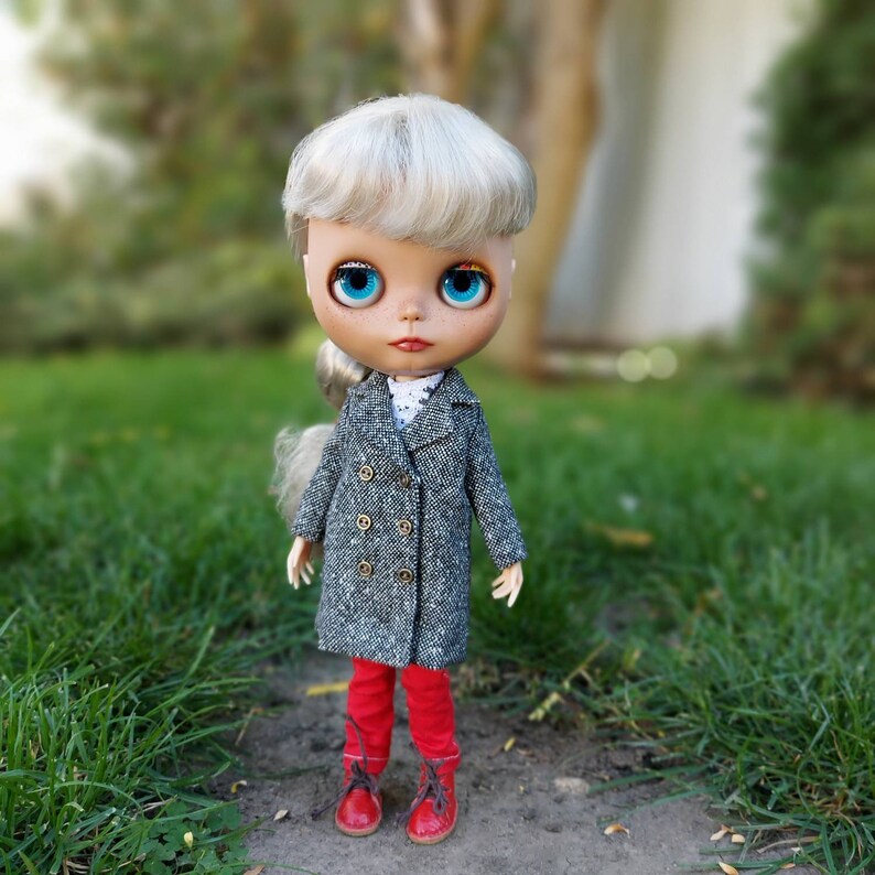 May include: A doll with blonde hair wearing a gray tweed coat, red pants, and red shoes. The doll has blue eyes and is standing on a grassy surface.