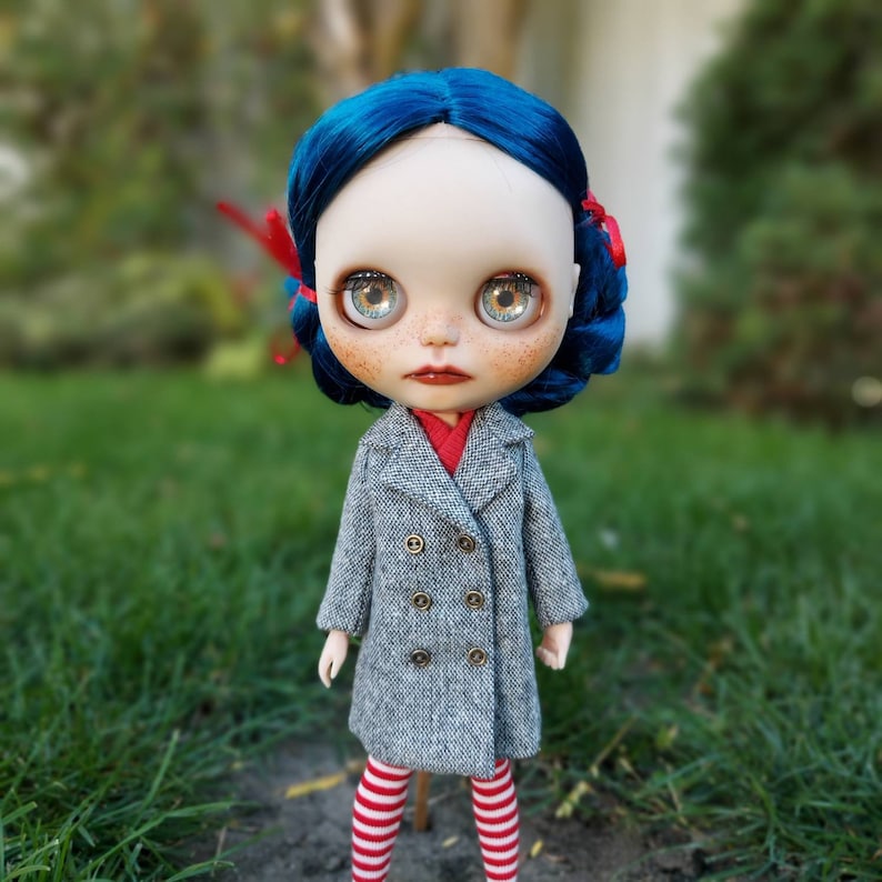 May include: A doll with blue hair, wearing a gray coat with a double breasted closure, red and white striped leggings, and red and white bows in her hair.