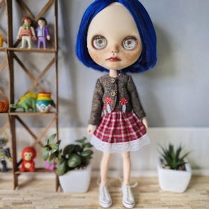 May include: A custom doll with blue hair, freckles, and large eyes. The doll wears a brown tweed jacket with mushroom appliqués, a red and white plaid skirt with a white lace hem, and white sneakers. The doll is standing in front of a shelf with toys.