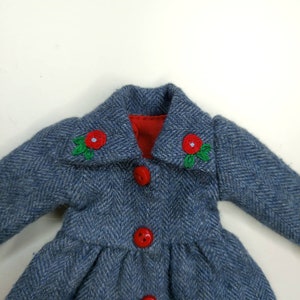 Puede incluir: Una chaqueta de tweed azul con forro rojo y botones rojos. El cuello tiene hojas bordadas verdes y flores bordadas rojas.