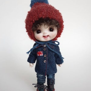 May include: A small doll wearing a red fluffy hat with a blue pom-pom, a blue denim jacket with a red, white and blue patch, blue denim trousers, and black boots.