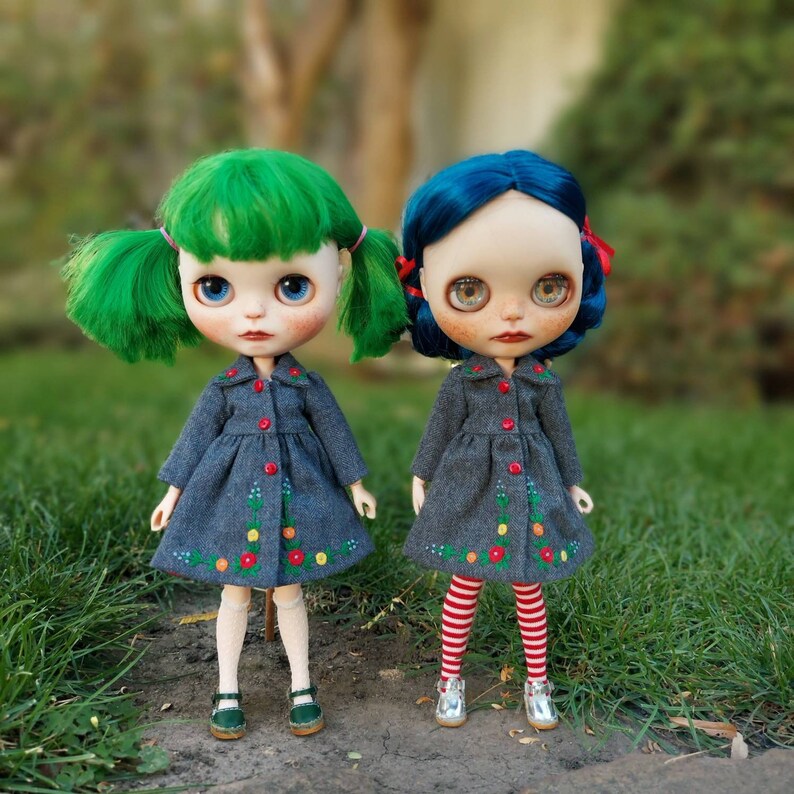 Puede incluir: Dos mu&ntilde;ecas con abrigos grises con bordados florales. Una mu&ntilde;eca tiene el pelo verde y la otra tiene el pelo azul. Ambas mu&ntilde;ecas llevan medias blancas y zapatos.