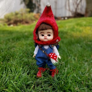 Può includere: Una piccola bambola con un cappello rosso con cappuccio e una salopette blu. La bambola tiene un fungo rosso e bianco. La bambola è in piedi in un'area erbosa.
