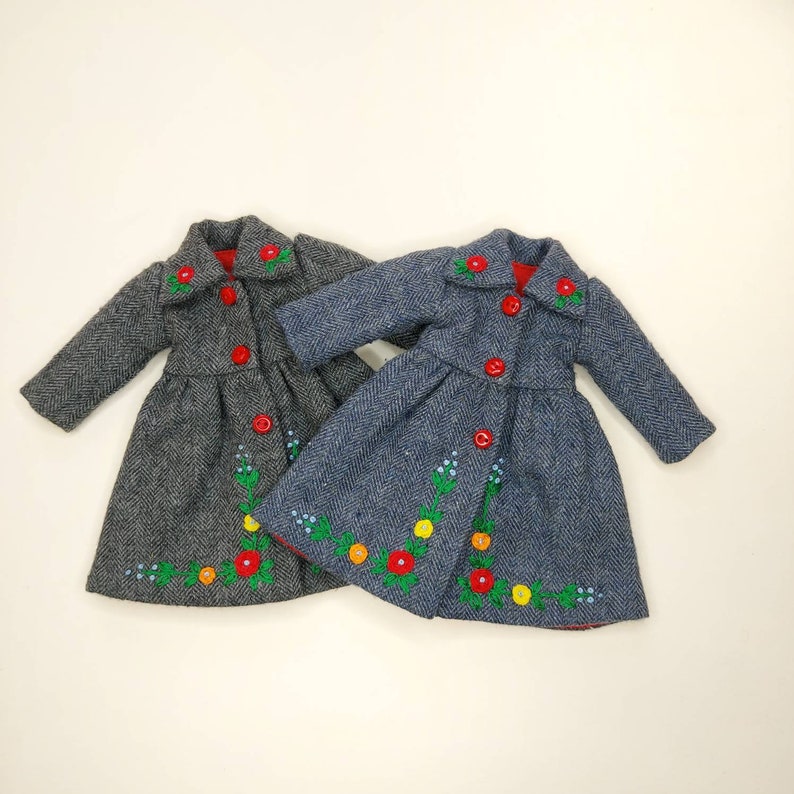Puede incluir: Dos abrigos de tama&ntilde;o de mu&ntilde;eca con detalles florales bordados. Un abrigo es gris con botones rojos y el otro es azul con botones rojos.