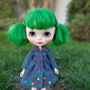 Puede incluir: Una mu&ntilde;eca con el pelo verde brillante, ojos azules y mejillas sonrosadas lleva un vestido de mezclilla azul con flores bordadas.