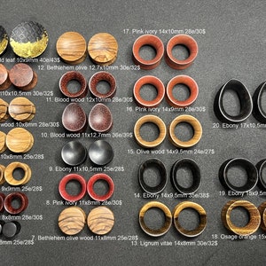 May include: A collection of 20 pairs of wooden ear gauges in various colours and wood types, including ebony, olive wood, blood wood, and pink ivory. The gauges are arranged in rows and labelled with their size and price.
