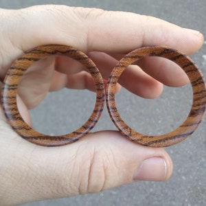 May include: Two wooden ear gauges with a striped pattern in shades of brown. The gauges are held in a hand.