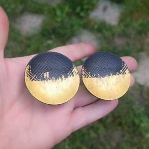 May include: Two round black and gold earrings with a geometric pattern. The earrings are made of metal and have a gold finish.