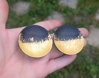 May include: Two round black and gold earrings with a geometric pattern. The earrings are made of metal and have a gold finish.