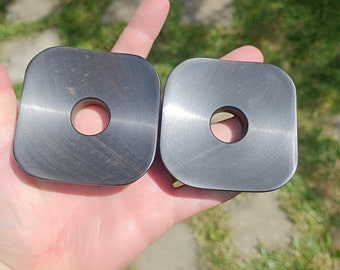 May include: Two black square-shaped wooden coasters with a round hole in the center. The coasters are slightly different sizes.