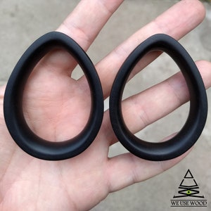 Puede incluir: Dos medidores de oreja de madera negra en forma de lágrima. Los medidores se sostienen en una mano.