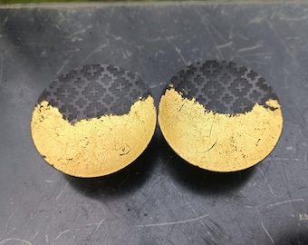 May include: Two round black coasters with a gold leaf design. The coasters have a textured surface and are half black and half gold.