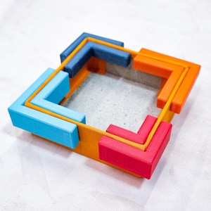 May include: A square-shaped box made of four interlocking corner pieces. The pieces are stacked in a pattern of blue, orange, and red.