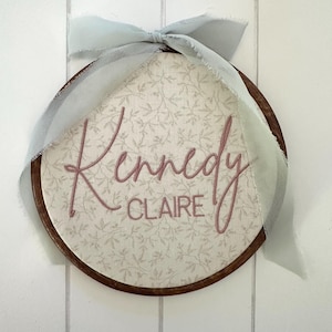 May include: A circular wooden embroidery hoop containing fabric with a light beige floral pattern. The name Kennedy Claire is embroidered in dusty rose thread across the center. A light blue frayed ribbon is tied in a bow at the top.