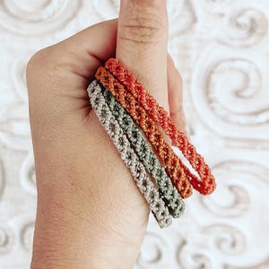 May include: Four macrame bracelets in shades of orange, green, and gray are held in a hand. The bracelets are made of a woven material and have a textured appearance. The background is a white, patterned surface.
