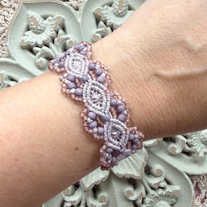 May include: A macrame bracelet featuring light gray knotted cord arranged in a repeating oval pattern, accented with matte lavender beads and small translucent pink seed beads woven along the edges to create a textured, decorative wrist accessory.