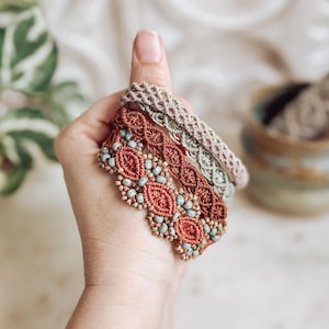 May include: Three macrame bracelets in shades of brown and green are held up by a hand. The bracelets feature intricate woven patterns and small, light blue beads. The bracelets are a bohemian style.