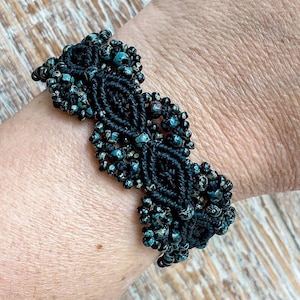 May include: Black macrame bracelet with turquoise and black beads. The bracelet features a central diamond pattern and beaded clusters. The intricate design is visible against the skin, showcasing the craftsmanship of the jewelry.