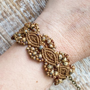 May include: A macrame bracelet crafted from brown cord, featuring an intricate woven pattern interspersed with small, multi-colored speckled beads in shades of tan, blue, and cream. The design includes repeating diamond-shaped knots.