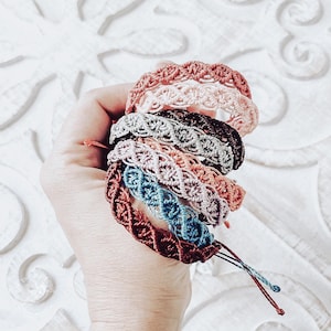 May include: A stack of handmade macrame bracelets in various colors, including pink, brown, blue, and gray. Each bracelet features a woven design and adjustable ties. The bracelets are held in a hand.