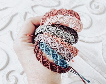 May include: A stack of handmade macrame bracelets in various colors, including pink, brown, blue, and gray. Each bracelet features a woven design and adjustable ties. The bracelets are held in a hand.