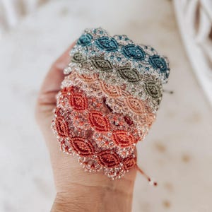 May include: A stack of four woven bracelets featuring intricate eye-shaped patterns in teal, olive green, light peach, and terracotta orange. Each bracelet is embellished with clear, translucent glass beads woven into the thread design.